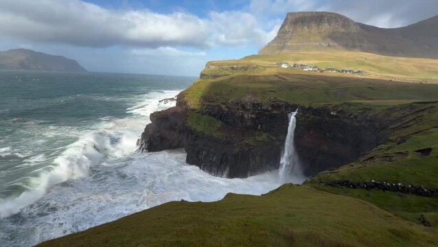 Mulafossur waterfall falling from grassy sea cliffs into crashing ocean waves in Faroe Islands