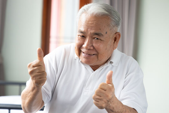 happy smiling elderly southeast asian man in his 70s giving two thumbs up with excitement and looking at the camera, home indoor scene