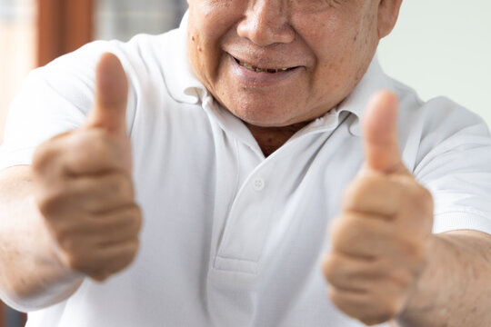 smiling mouth close up shot of happy elderly southeast asian man in his 70s giving two thumbs up, home indoor scene with natural window light