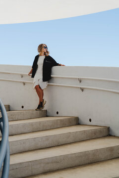 Woman in sunglasses and black jacket leans on a curved parapet at the top of a wide outdoor staircase under clear sky. Minimalist architecture and urban fashion for travel and lifestyle branding.