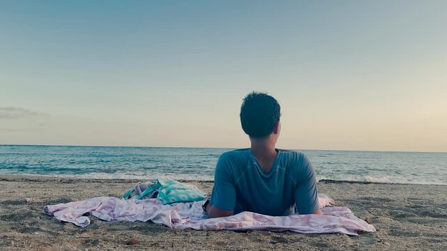 Man lying on the sand while gazing at the Mediterranean horizon, changing positions during a peaceful sunset by the beach