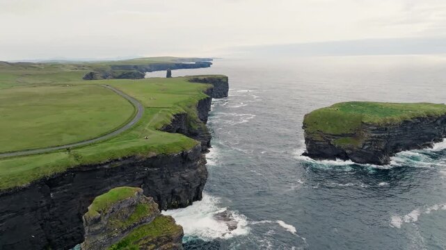 An offshore sea stack rises beside layered sea cliffs on the Kilkee coast in County Clare, Ireland, viewed from above with Atlantic surf and coastal pasture.