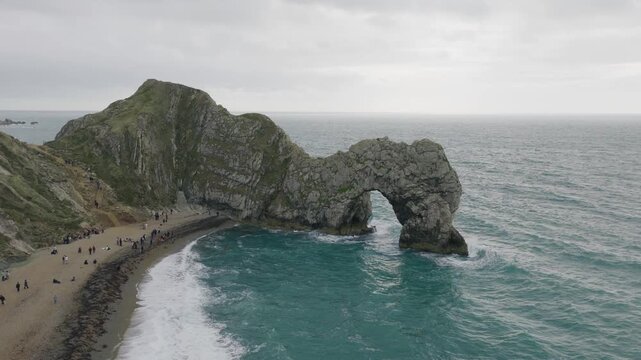 Front aerial view of Durdle Door in Dorset showing the iconic limestone sea arch beach visitors turquoise water and waves along the Jurassic Coast