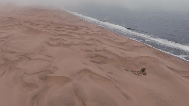 A breathtaking cinematic aerial shot showcasing the vast sand dunes of the Namib Desert meeting the crashing waves of the Atlantic Ocean under a misty sky. Ideal for travel and sustainability content.