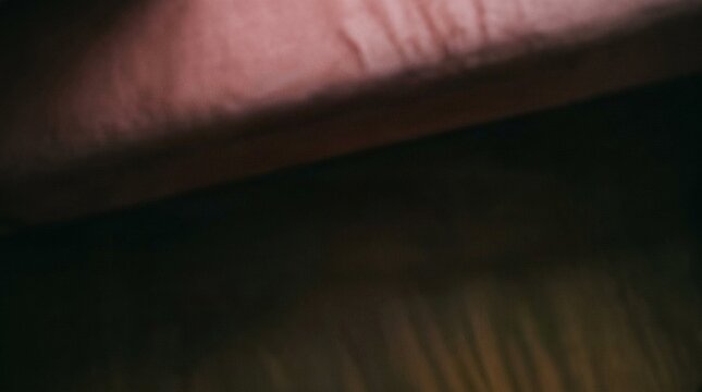 Dark abstract photograph featuring smooth textured dusty pink fabric edge contrasting deeply against a mysterious blurry dark background during indoor low light photographic conditions