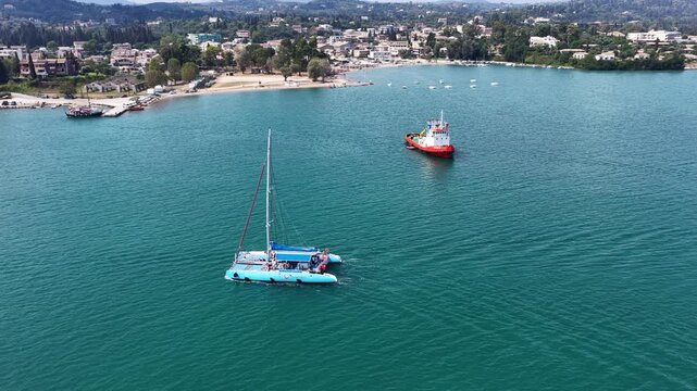 Wide aerial view of a blue catamaran sailing past a red tugboat in the turquoise coastal bay of Corfu island, Greece