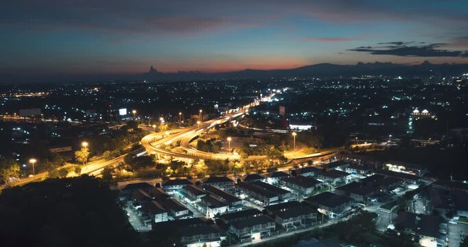 Timelapse aerial view of traffic flowing through Don Chan Intersection in Chiang Mai, Thailand at twilight with bright road light trails, urban neighborhoods, far mountains and a colorful evening sky.