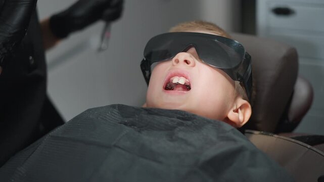 Close up of young boy lying on dental chair wearing protective glasses during orthodontic procedure. Dental professional in black gloves prepares instrument near open mouth focusing