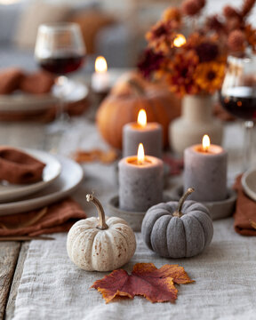 Cozy autumn table bathed in candlelight. Candles flicker beside rustic pumpkins and leaves. Wine glasses hold deep red wine, softly blurred. Plates rest on linen, surrounded by warm tones