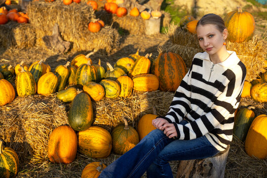 Young woman sitting among colorful pumpkins and hay bales in warm autumn sunlight, rustic harvest setting with abundant seasonal gourds.