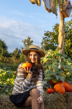 Young woman in straw hat holding orange pumpkin while sitting on hay in autumn garden, warm sunlight creates a cozy seasonal harvest atmosphere.