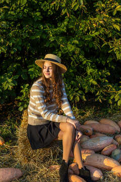 Young woman in straw hat sitting on hay bale beside pumpkins in warm autumn sunlight, peaceful seasonal portrait surrounded by lush green foliage.