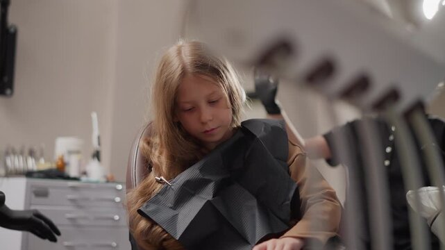 Close up of young girl sitting in dental chair with black bib receiving orthodontic treatment from professional wearing gloves. Dental tools and equipment visible on counter behind her. Soft