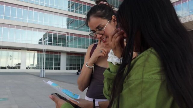 Female friends using a paper map and smartphone for directions, happily navigating a modern city while enjoying their vacation trip and exploring new urban destinations
