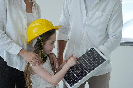 Happy young family with little daughter in a toy hard hat let the girl in a toy helmet touch the photovoltaic cells up close inside their new energy efficient home, candid real moment