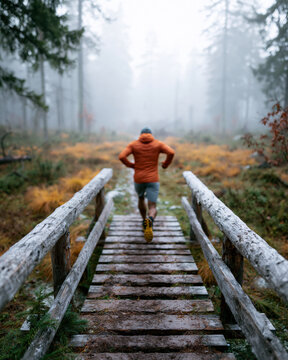 Mist wraps tall trees in quiet haze. Runner strides forward on weathered wooden bridge. Orange jacket contrasts muted forest tones. Footsteps echo softly on damp planks. Mood suggests determination