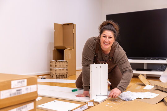 Adult woman kneeling barefoot on a polished wooden floor, assembling flat‑pack furniture