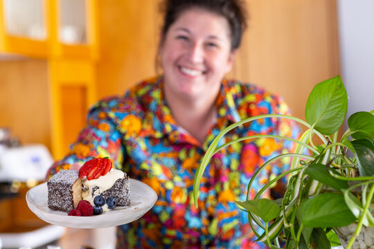 Home baker handing a plated chocolate-coated sponge cake with whipped cream and fresh berries