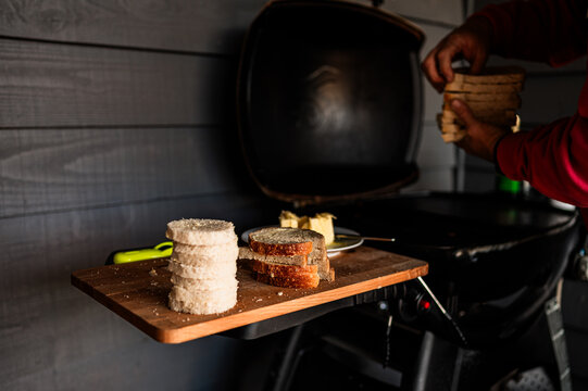 Slices of bread are ready on a wooden board while someone arranges more bread on a gas barbecue