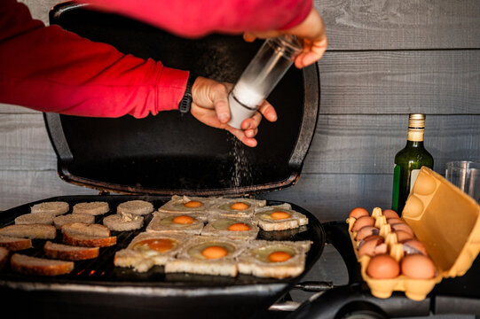 A person seasons eggs and bread with salt on a gas barbecue while preparing breakfast outdoors