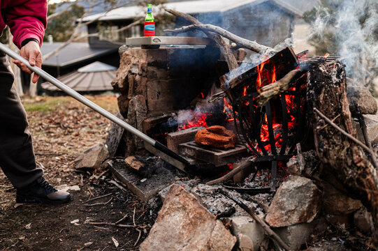 A person tending a fire pit using a long metal tool to adjust burning logs and embers