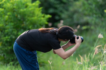 An LGBTQ woman bends forward to photograph wild grasses outdoors against lush green background © NongEngEng