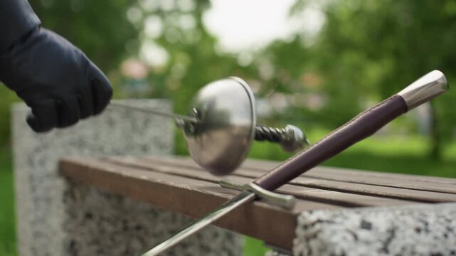 Close up of wooden bench with metal swords placed on it in park setting. Background shows green trees and soft daylight creating calm atmosphere. Bench made of wood slats supported by stone