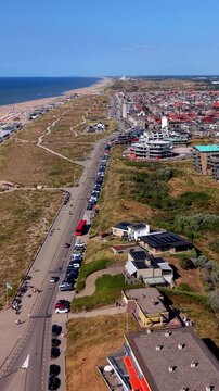 Vertical aerial drone clip of coastal boulevard, North Sea beach, sand dunes, red bus, and seaside town in Katwijk aan Zee, South Holland, Netherlands on sunny summer day