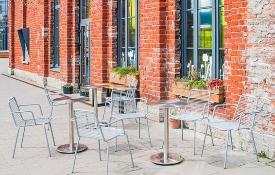 Quaint outdoor caf&eacute; setting with metal chairs and tables against a rustic red brick building, adorned with window planters.