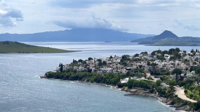 A elevated scenic view of Bitez in Bodrum, Turkey, showing white coastal houses, blue sea waters, an island, and mountain ridges under a cloudy sky.