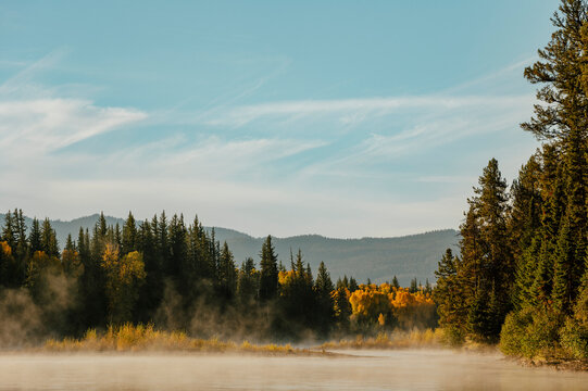 Morning mist drifts above a calm river bordered by autumn forest.