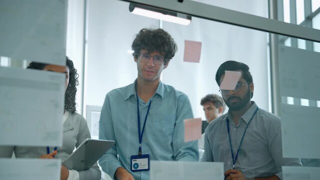 Engineering team collaborating around a glass board with digital project information, sticky notes and tablet devices in a modern office.
