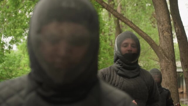 Close up of medieval reenactors in black padded armor walking through forested path. group maintains formation with lead figure slightly ahead and others following behind. Sunlight filters