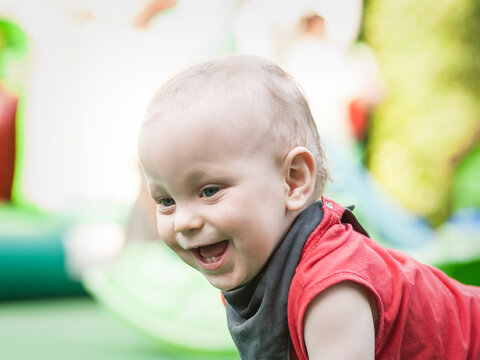 A cheerful toddler crawls on a green play mat, beaming with joy and curiosity. The vibrant surroundings include fun play structures, creating a lively atmosphere perfect for children.