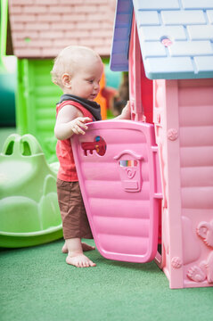 A cheerful toddler, barefoot and curious, opens the door of a pink playhouse while surrounded by bright play structures. This lively scene unfolds in a sunny playground, inviting imagination.