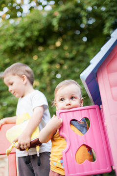 Two young children enjoy their time outdoors, engaging with a bright pink playhouse and colorful toys in a lush green backyard. Their laughter fills the air under a clear sky.