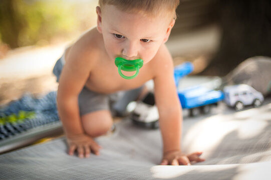 A cheerful toddler crawls across a soft blanket, surrounded by colorful toy vehicles. The sun shines brightly, creating a playful atmosphere in a backyard or park setting.