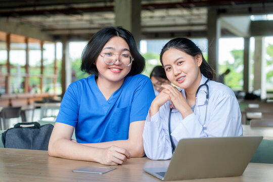 Asian medical students or nursing interns using laptop in university library, happy young healthcare workers in scrubs and lab coat studying together online