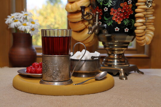 On the table, against the backdrop of the autumn landscape outside the window, there is a glass of black tea in a cup holder and a tray with sugar and sweets, and next to it is a samovar with bagels. 