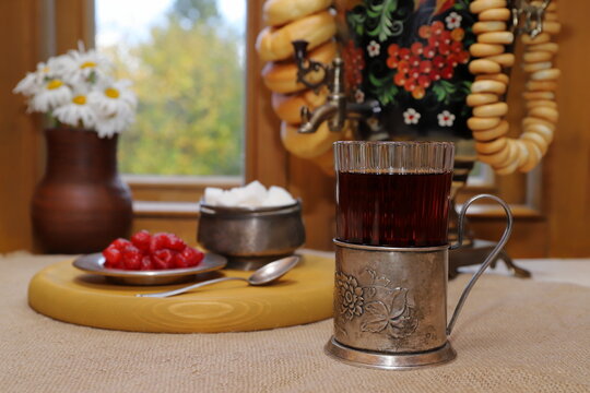 On the table, against the backdrop of the autumn landscape outside the window, there is a glass of black tea in a cup holder and a tray with sugar and sweets, and next to it is a samovar with bagels. 