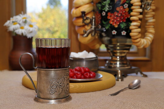 On the table, against the backdrop of the autumn landscape outside the window, there is a glass of black tea in a cup holder and a tray with sugar and sweets, and next to it is a samovar with bagels. 
