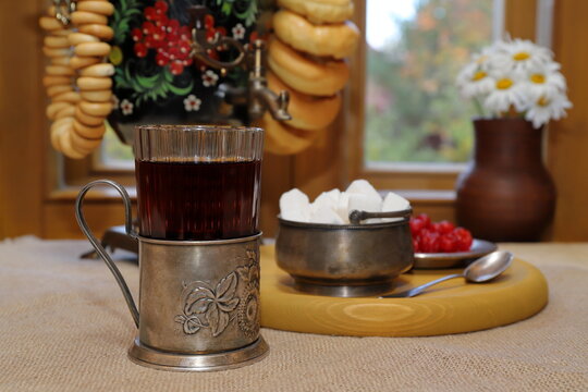 On the table, against the backdrop of the autumn landscape outside the window, there is a glass of black tea in a cup holder and a tray with sugar and sweets, and next to it is a samovar with bagels. 