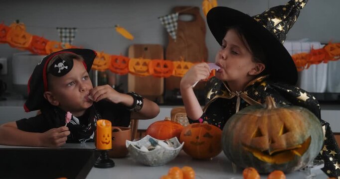 A medium shot of children in Halloween costumes decorating their home, carving a pumpkin, and eating gummy candies. They are playing together and celebrating Halloween in the kitchen. Trick or treat.