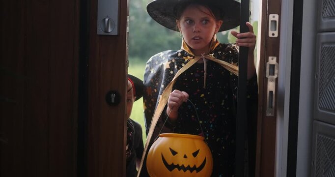 A medium shot of children in Halloween costumes opening doors and holding out pumpkin shaped baskets while saying Trick or treat. Celebrating Halloween with children and collecting candy.