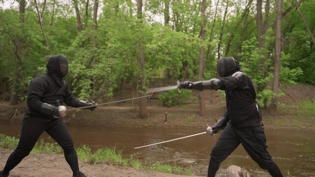 Two men in black fencing gear engage with swords on dirt path beside river surrounded by dense green trees. Their movements are precise as they parry and thrust demonstrating skillful
