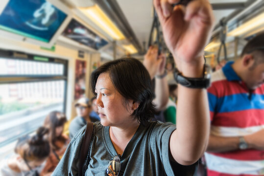 An Asian woman holds an overhead strap looks out the window while traveling other passengers crowded train carriage