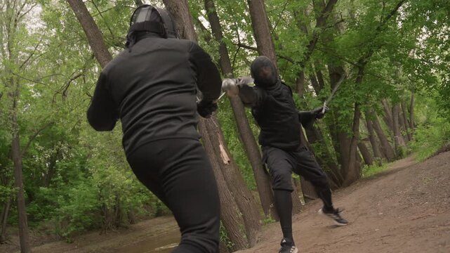 Two men in black martial arts gear engage in sword fight along forest path demonstrating precise movements and techniques. scene features dense green foliage surrounding action with natural