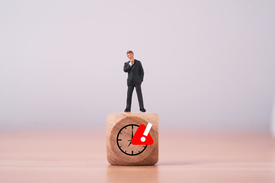 Businessman standing on a wooden clock block with red warning symbol for time pressure from deadline risk, urgent decisions, time management challenges and business uncertainty in workplace.