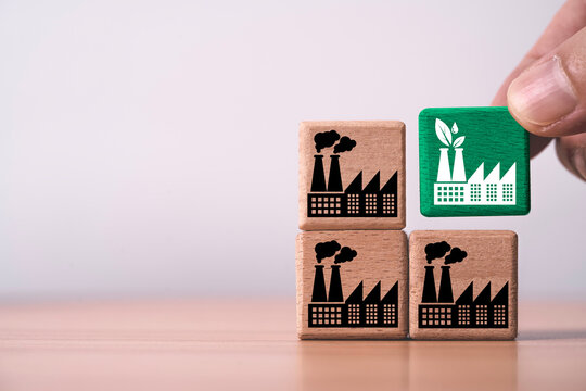 Hand placing a green factory symbol among wooden blocks polluting industries representing sustainable manufacturing, industrial decarbonization to clean technology and green energy transition.