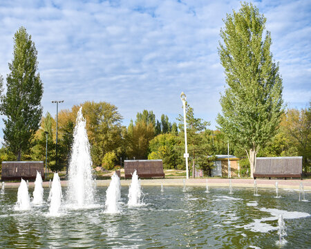 A large round fountain in a public park with high central jets and cascading water streams surrounded by autumn trees under a blue sky